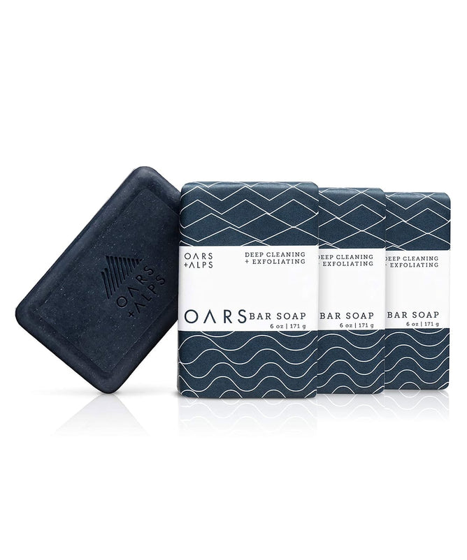 Oars + Alps Exfoliating Bar Soaps are in dark packaging with wave designs. An unwrapped charcoal soap at the front displays the brands embossed logo, promising a deep exfoliation experience.