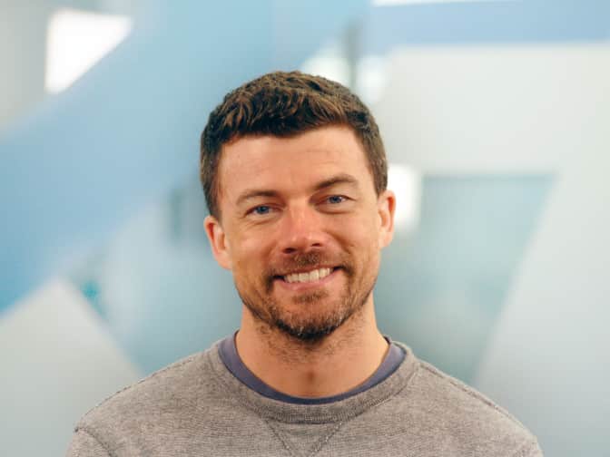 Image of white male with brown hair and facial scruff in grey shirt