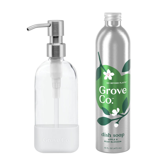 A transparent glass soap dispenser with a metal pump is beside a silver bottle labeled Grove Co. The Dish Soap Starter Set, featuring green leaves and white blossoms, delivers an apple and pear blossom scent. It helps reduce plastic waste, adding an eco-friendly touch to your kitchen.