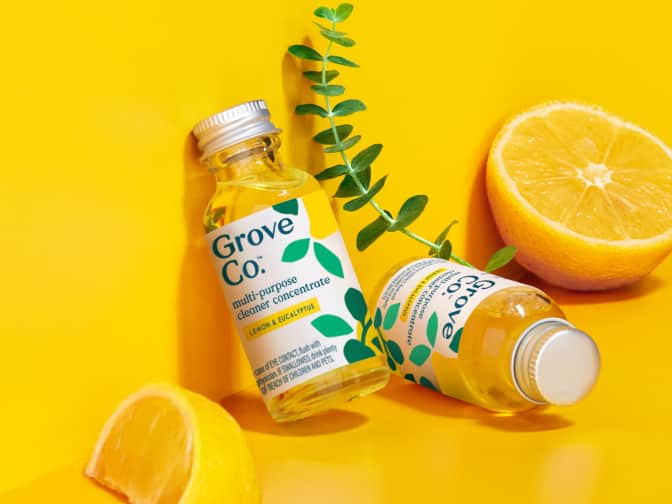 A Grove Co. glass dish soap dispenser lying next to a bottle of Grove Co. Ultimate Dish soap on a brightly colored yellow background with lemons and eucalyptus leaves scattered around.