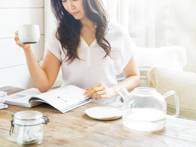 woman reading a book while holding a mug of coffee by a window