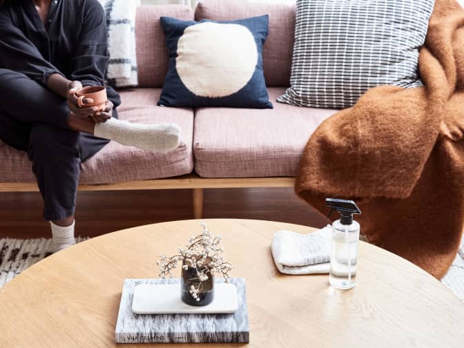Image of person sitting on a couch with a cup of coffee and coffee table with spray and cloth
