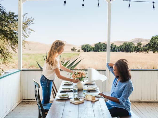 Young girl fixing flower centerpiece on a plated patio picnic table with Black woman sitting down across