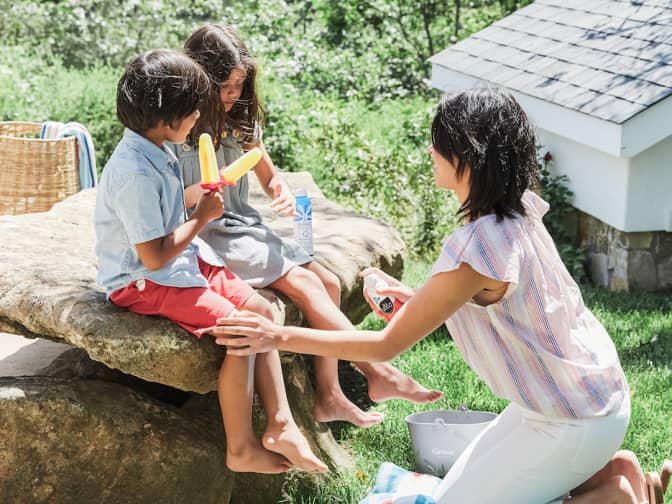 Image of woman outside putting sun product on her kids legs while they eat popsicles