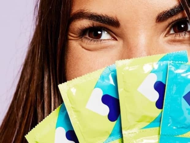 Person with brown hair holding several packages of condoms in front of their face, only showing their eyes