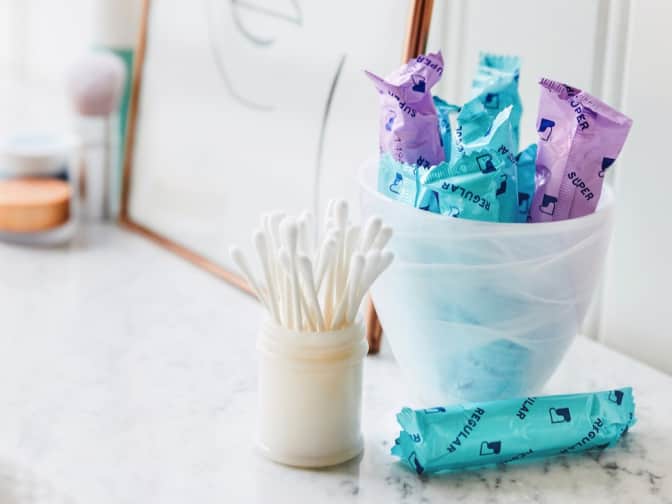 clear plastic container holding sustain tampons wrapped in blue and purple wrappers next to cup of q-tips on a vanity