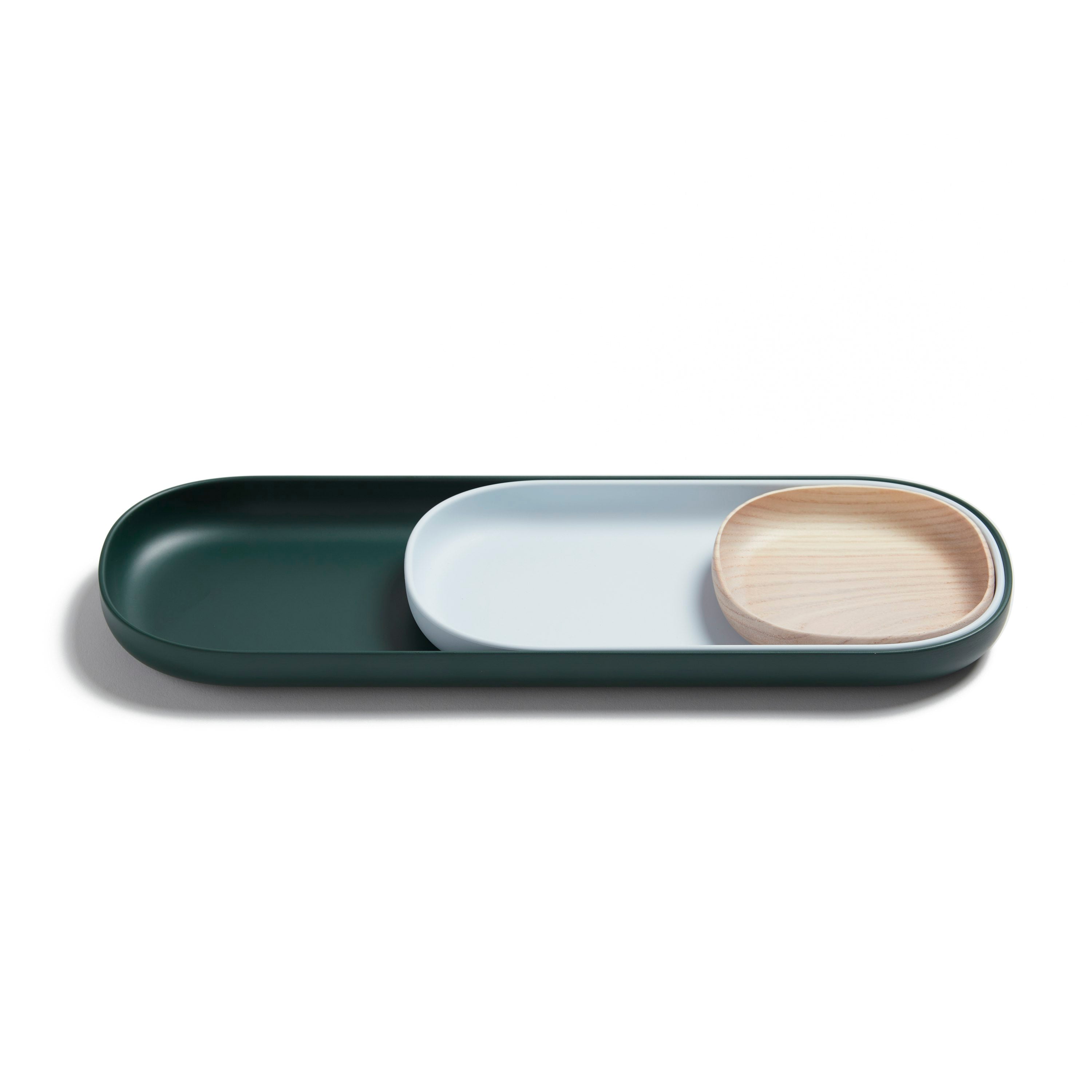 The Nesting Tray Set by Open Spaces features three elegantly arranged trays: a large dark green tray, a medium white oval tray, and a small round light wood tray, all boasting a minimalist design on a pristine white surface.