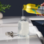 Someone refills a clear sink dispenser with Grove Co.s Hydrating Hand Soap - 24oz, packaged in a yellow bottle featuring a green leaf design. A tap and plant enhance the tranquil ambiance in the blurred background.
